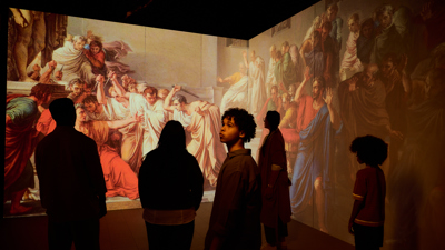 A group of people looking at scenes depicting Caesars murder projected on wall screens in ROME: Empire, Power, People exhibition in Melbourne Museum. 