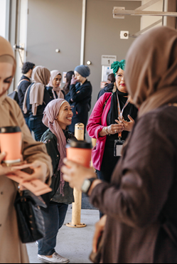 Many Muslim women, wearing a variety of colourful hijabs, stand in groups and network over coffee. 