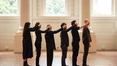 Six performers standing side-by-side, each reaching forward to cover another performer’s eyes