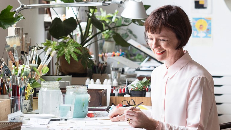Anna Walker writing and illustrating at her desk covered in paints and brushes and house plants.