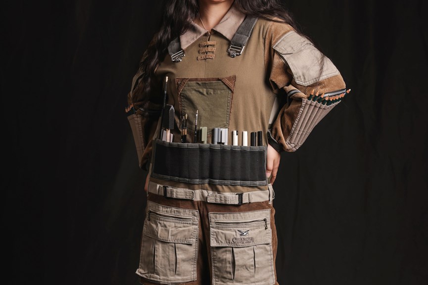 Person wearing an earth-toned jumpsuit with corduroy patches. Several pockets on the front and arms of the jumpsuit hold art materials, including pencils, pens and paintbrushes.