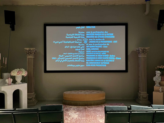 A wall-mounted screen displays film credits in Arabic and French. Under the screen is a beige oval ottoman, and white Greek columns, flowers and white busts on either side. In the foreground is the first row of audience seating with an aisle in the middle.