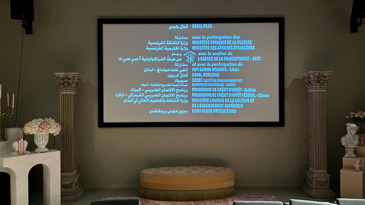 A wall-mounted screen displays film credits in Arabic and French. Under the screen is a beige oval ottoman, and white Greek columns, flowers and white busts on either side. In the foreground is the first row of audience seating with an aisle in the middle.