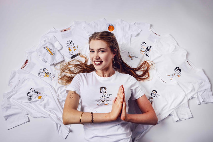 Smiling person lying on a white background with arms in a prayer position, surrounded by white T-shirts with hand-drawn illustrations.