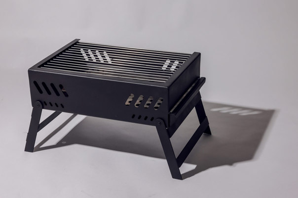 Black metal portable barbecue with slatted grilling surface and ventilation slots on sides, supported by foldable legs.
