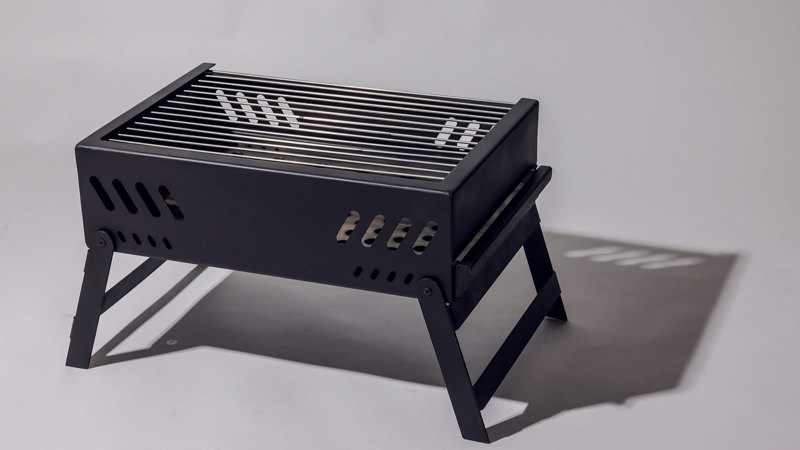 Black metal portable barbecue with slatted grilling surface and ventilation slots on sides, supported by foldable legs.