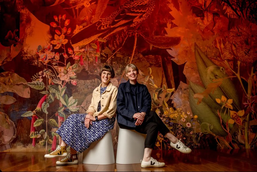 Two women seated on stools in front of a large red collage with leaves, birds, flowers and photograph snippets.  