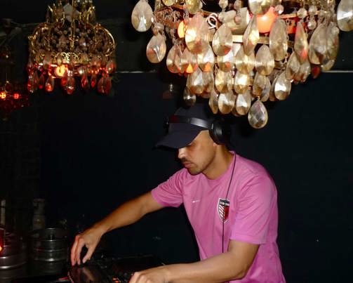 A spotlighted side photo of a man in a black cap and pink t-shirt, wearing black headphones and operating a DJ table in front of him. Above his head are two gold and crystal chandeliers, reflecting the camera's flash. 