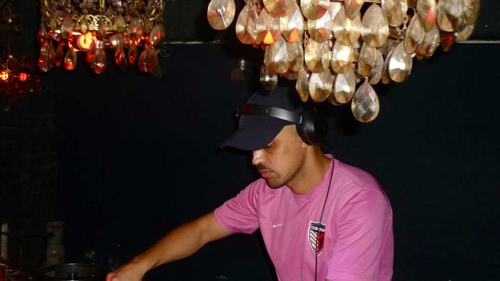A spotlighted side photo of a man in a black cap and pink t-shirt, wearing black headphones and operating a DJ table in front of him. Above his head are two gold and crystal chandeliers, reflecting the camera's flash. 