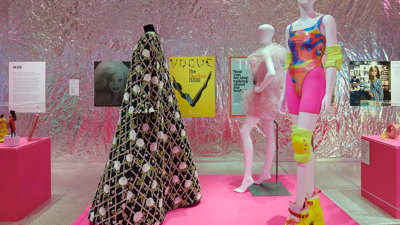 An image of three mannequins with movie costumes. The wall in the background is covered is lined with artwork and covered with metallic foil as wallpaper.