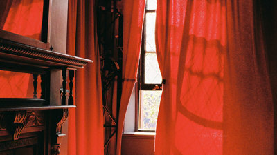 A red sunlit room with light coming through a crack in the curtains and falling on a bed and furniture. 