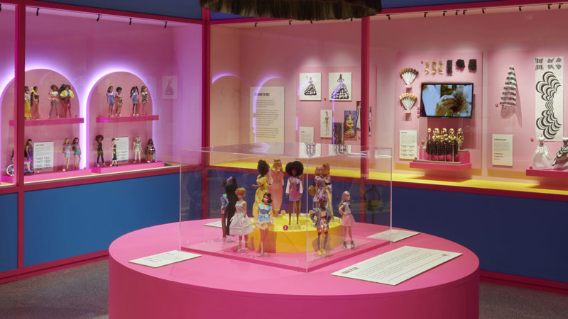 Barbies in a square glass display case on a circular table in a room lined with more displays of barbie showing designs and digital display screens.