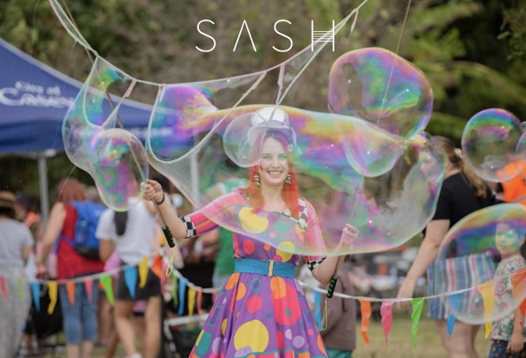 A woman with a colourful spotted dress smiles while making huge bubbles.