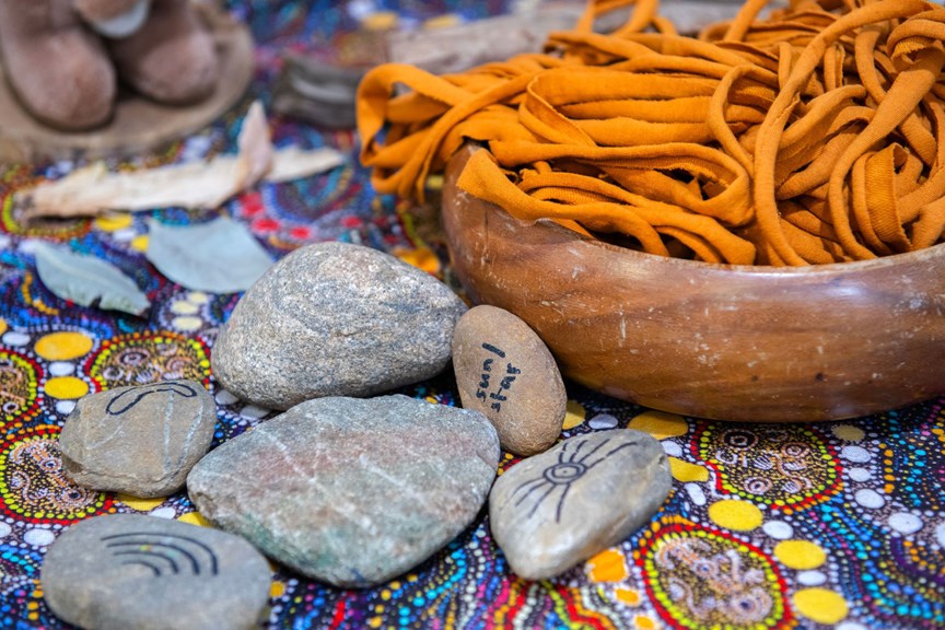 A wooden bowl containing orange rope sits beside illustrated river stones on a surface decorated in indigenous dot painting art.