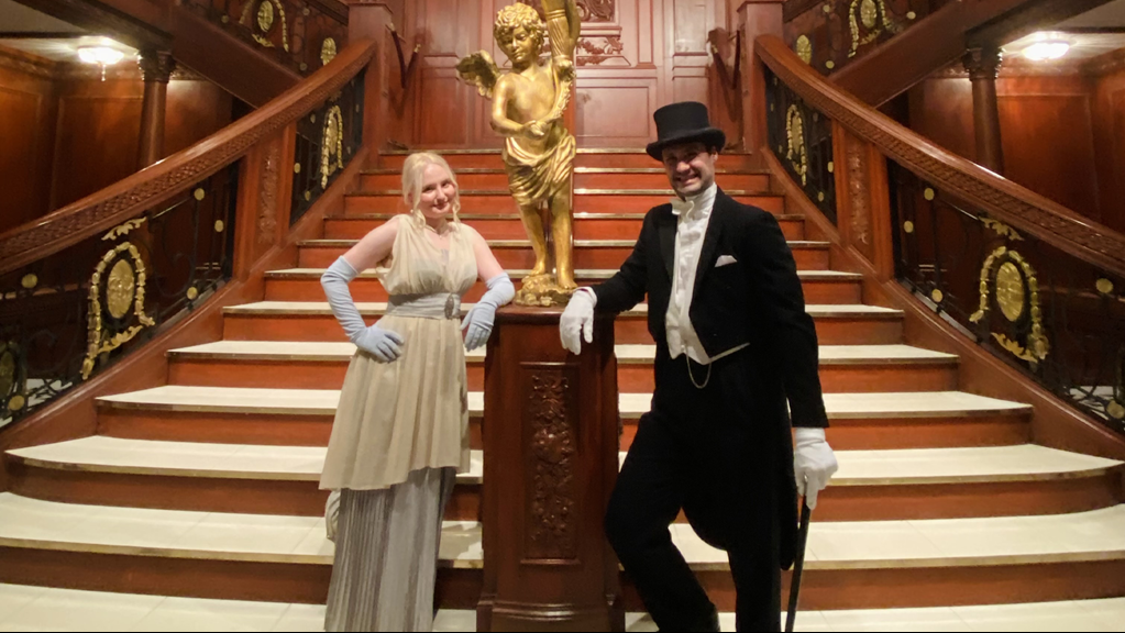 A man and woman in a 1914 period dinner party attire pose on the grand staircase of the Titanic