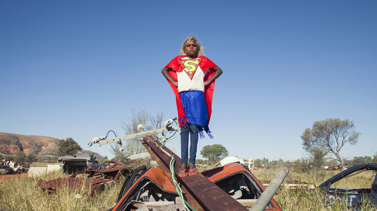 A child wears a hand-made Supermen-like costume and stands on a crushed car and electric pole. 