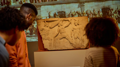 Visitors viewing a Roman stone relief of Gladiators in action, with a large projected crowd scene behind it. 