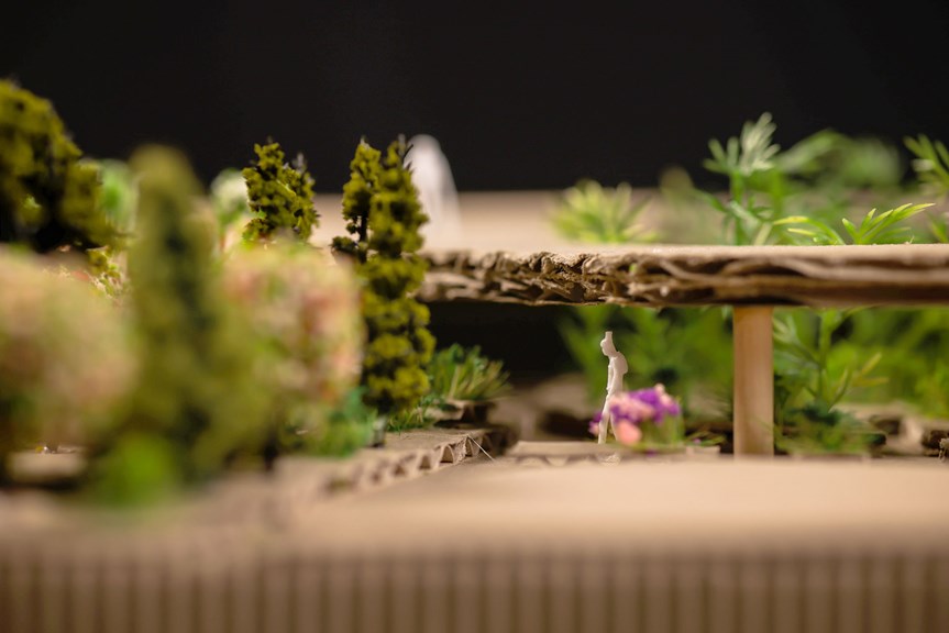 Close-up view of an architectural model with trees, flowers, and a scale model white figure in the middle of frame.