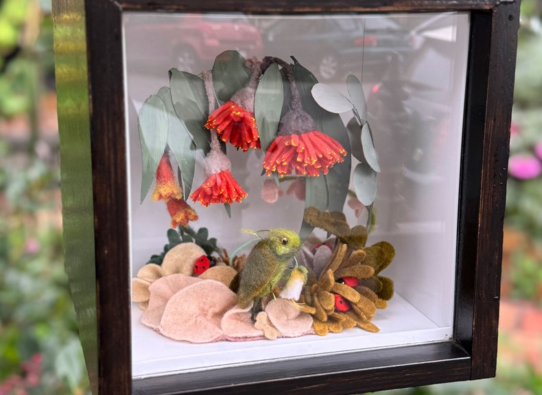 Wattle flowers made from felt and real gum leaves sit above a green felt fantasy creature. This scene is a diorama behind glass in a wooden box.