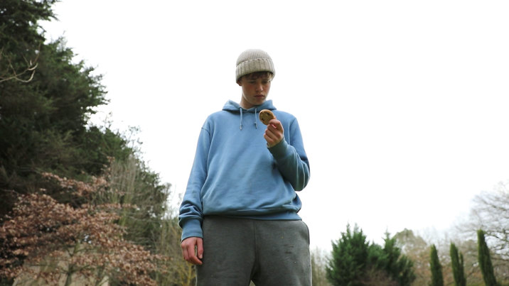 Film still showing a teenaged boy in a beige beanie and blue hoodie holding a chocolate-chip cookie, against a backdrop of trees and an overcast sky.