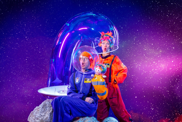 Two performers in bright orange and blue space suits with bubble helmets sit on an asteroid as it zooms through space.