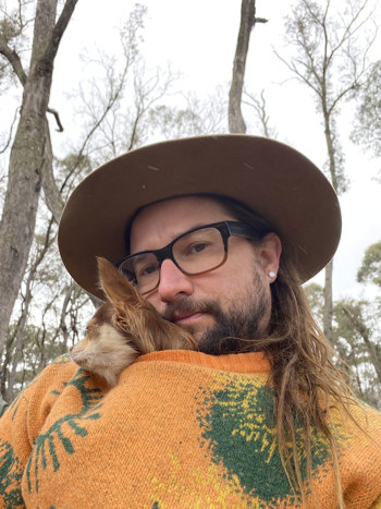 Mark Free with a wide brim hat, glassed, beard, long brown hair and a dog poking its head through a orange jumper he is wearing.