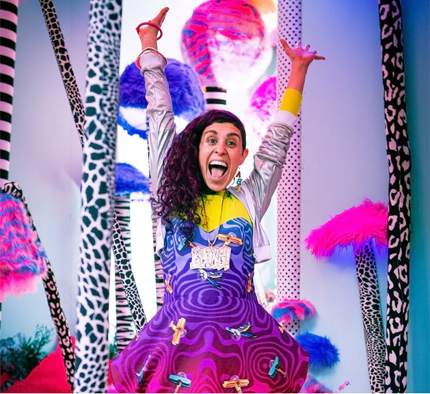 Femcee, wearing a colourful jacket, smiling and raising her hands.