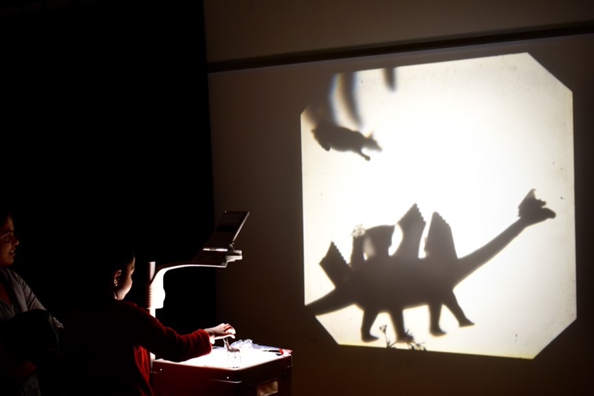 A young child plays with objects laid out on a projector, as a large dinosaur shadow is projected onto a white wall in front of them. 