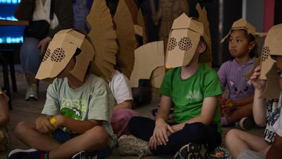 Children sit wearing Roman-style helmets with cardboard face guards.