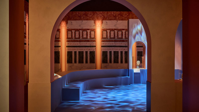 Arched gallery space inspired by a Roman bathhouse, with columns, projected light patterns, and a small sculpture in an alcove in ROME: Empire, Power, People exhibition in Melbourne Museum.. 