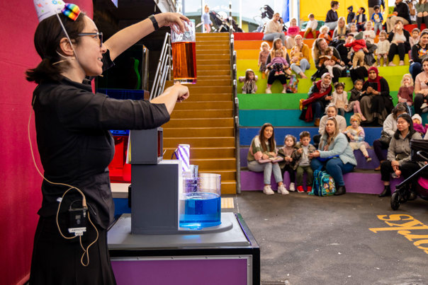 A presenter holds up a container with orange-coloured water above a container filled blue-coloured water.  