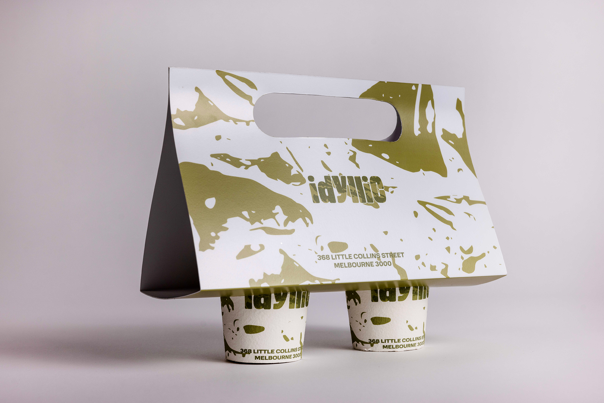 Coffee cup carrier with a gold and white splattered design, holding two branded takeaway cups from Idyllic Cafe.