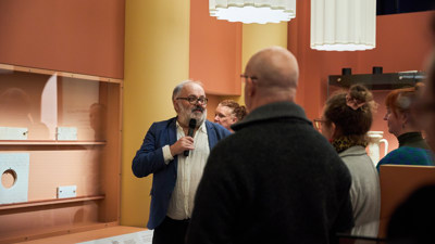 Man with grey hair, beard and glasses with a microphone leading a tour around the Rome exhibition at Melbourne Museum. Audience in the background.