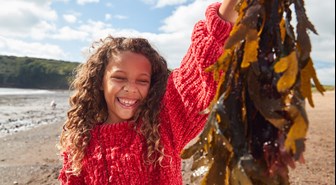 Smiling young girl with curly hair and red jumper holds up a large handful of dangling seaweed.