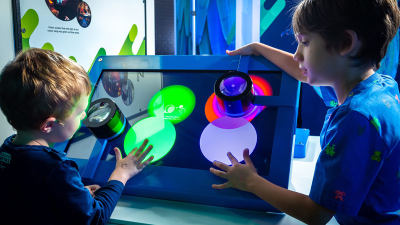 Two children interact with a colourful, hands‑on light exhibit. They place their hands on a large illuminated screen where overlapping circles of green, blue, purple, and red light create bright blended patterns. The display is framed by a blue structure, with mounted lights projecting the colours onto the surface. 
