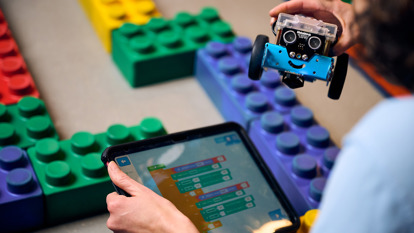 A girl looks at an m-bot robot, she holds an iPad with block programming in her lap.