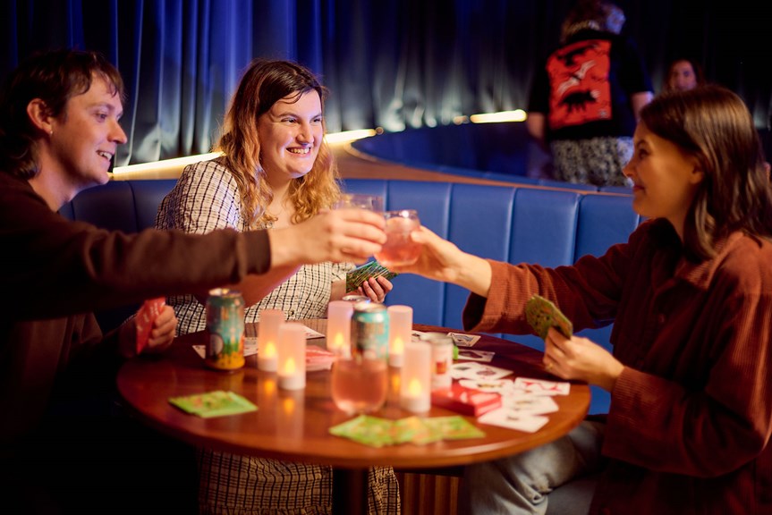 Three adults drinking, playing cards and eating jaffles during Adult Museum Sleepovers at Melbourne Museum.