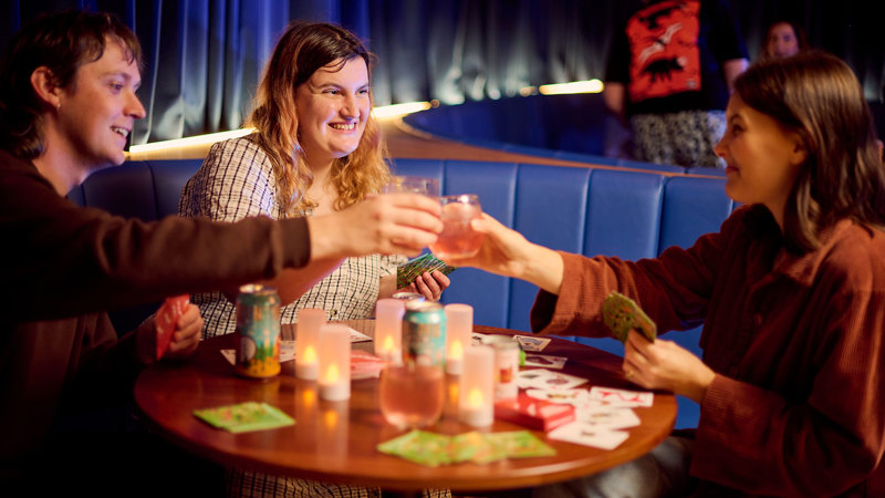 Three adults drinking, playing cards and eating jaffles during Adult Museum Sleepovers at Melbourne Museum.