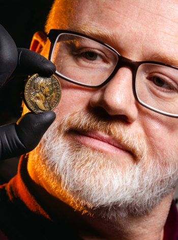 Close up of a bearded man holding a  roman coin with gloves.