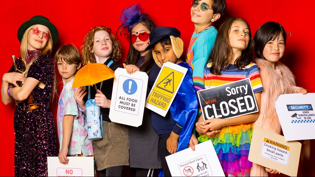 Children in dress up costumes, standing in front of a red wall holding signs like ‘tipping hazard’. 