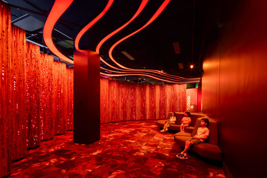 Three children take a rest in a room with red light illuminating a panelled wall display. 