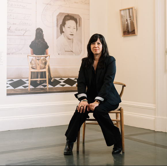 Pia Johnson, wearing a black suit, sits in front of a photographic exhibition of her self-portraits. 