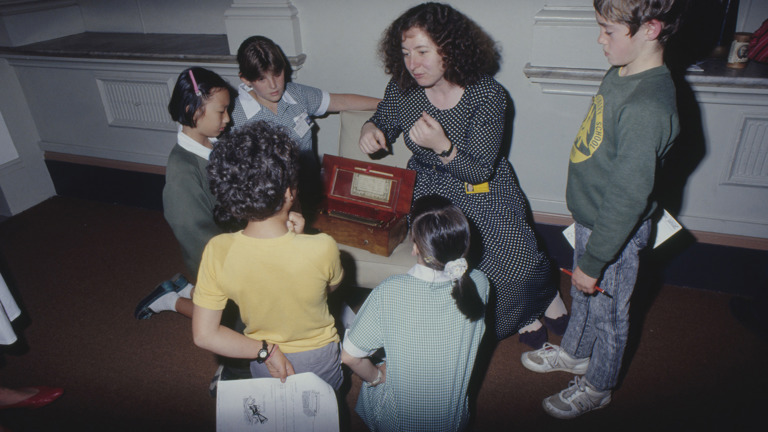 A teacher speaks to a group of children crouching in a circle around a small wooden chest.