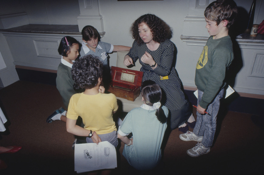 A teacher speaks to a group of children crouching in a circle around a small wooden chest.