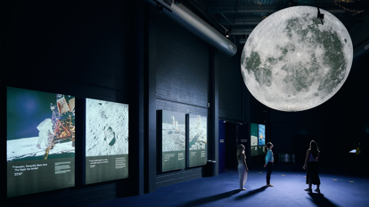 Three people stand in the Planetarium foyer in Scienceworks, looking at a hanging sculpture of the Moon. The Moon is detailed and centrally lit, so that it glows.