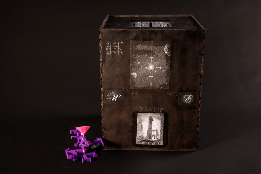 Black box decorated with lettering and illustrations. A 3D printed purple geometric key sits next to the box.