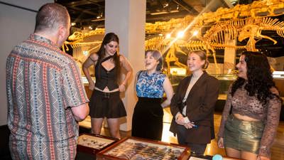  Visitors to Nocturnal x The Fashion Edition, speak to the entomology collection museum specialist in front of a table of specimen display cases, with dinosaur skeletons behind them.