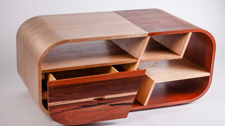 Rectangular coffee table in mixed wood tones with curved edges, enclosed sides and open compartments segmented by angled dividers. One front drawer is partially opened.