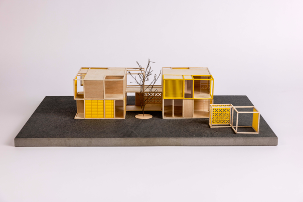 Wooden architectural model of modular housing with yellow accents, set on a dark grey base.