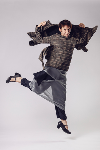 Person in a brown tweed oversized top and sheer grey trousers jumps with arms extended.